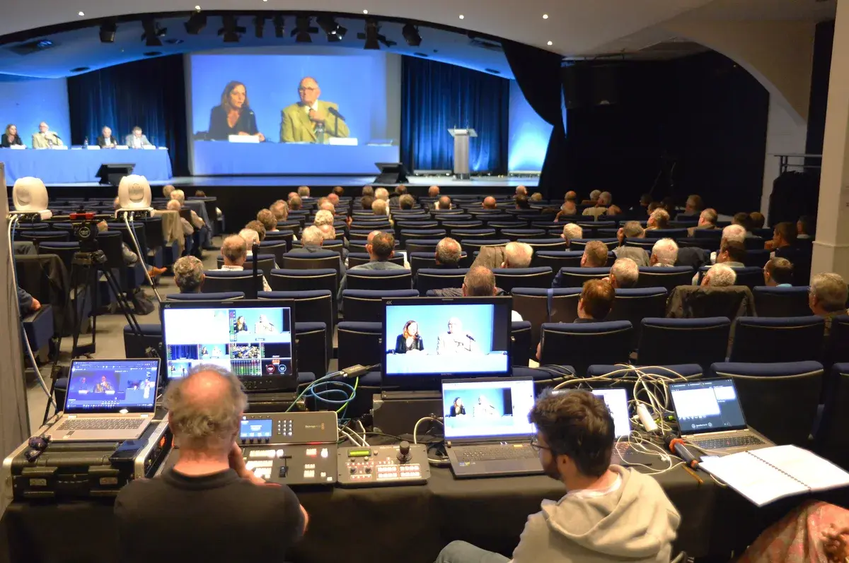 Video control station set up at the back of an auditorium during a meeting relayed on the main screen